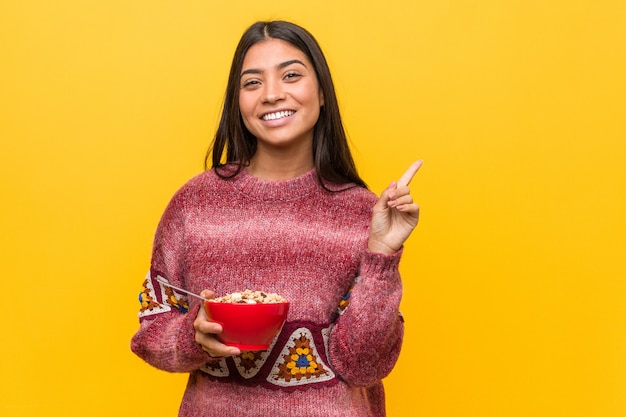 Giovane donna araba che tiene una ciotola di cereali che sorride allegramente indicando con l'indice di distanza.