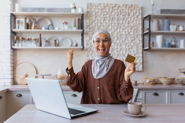 Giovane donna araba seduta a casa in cucina utilizzando un computer portatile utilizzando una carta di credito felice con la mano