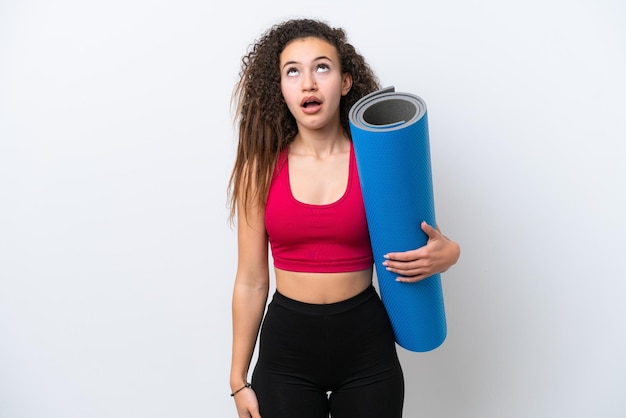 Giovane donna araba sportiva che va a lezioni di yoga mentre tiene un tappetino isolato su sfondo bianco guardando in alto e con espressione sorpresa