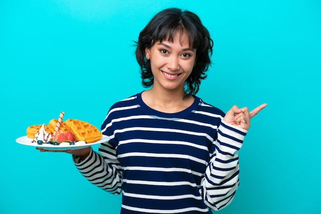 Giovane donna argentina che tiene i waffle isolati su sfondo blu che punta il dito di lato