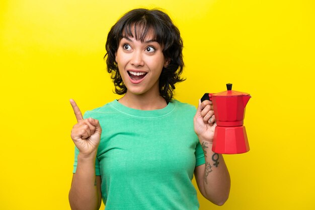 Giovane donna argentina che tiene la caffettiera isolata su sfondo giallo che intende realizzare la soluzione mentre solleva un dito