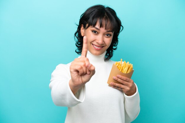 Giovane donna argentina che tiene patatine fritte isolate su sfondo blu che mostra e alza un dito