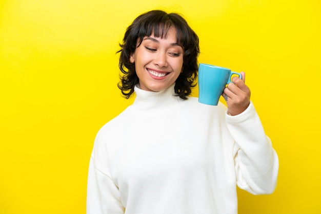 Giovane donna argentina che tiene una tazza di caffè isolata su sfondo giallo con felice espressione