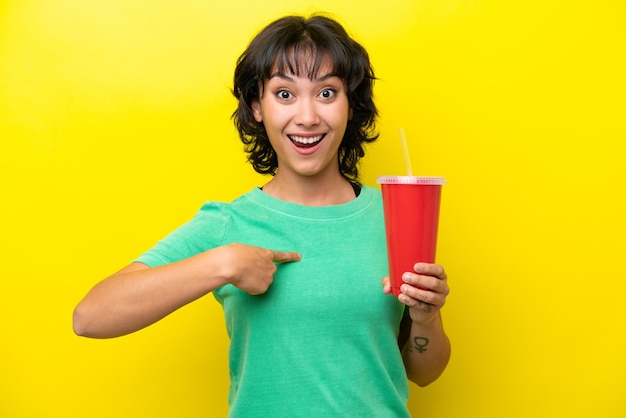 Giovane donna argentina in possesso di una soda isolata su sfondo giallo con espressione facciale a sorpresa