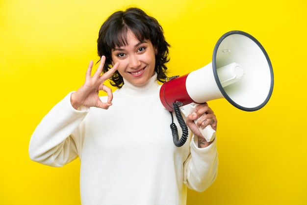 Giovane donna argentina isolata su sfondo giallo che tiene un megafono e mostra il segno giusto con le dita