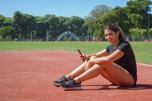 Giovane donna argentina seduta in pista utilizzando il cellulare e guardando la telecamera