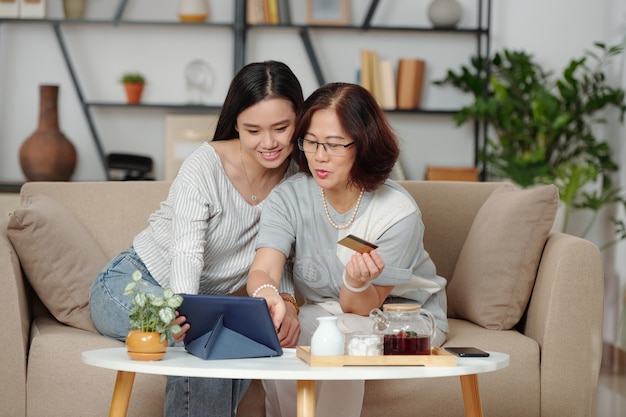 Giovane donna asiatica abbastanza sorridente che aiuta la madre a fare acquisti online e a pagare gli acquisti quando sono seduti sul divano in soggiorno e bevono tè