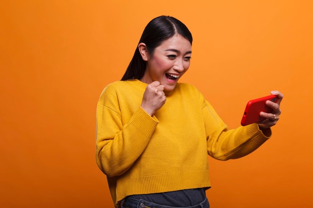 Giovane donna asiatica adulta che celebra la vittoria del gioco sul dispositivo smartphone. Persona positiva giocosa felice che indossa un maglione giallo che celebra il gioco vincente del telefono cellulare mentre è su sfondo arancione.