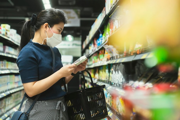 Giovane donna asiatica adulta che indossa una maschera protettiva per il viso scegliendo a mano un prodotto fresco o un prodotto di consumo nel centro commerciale del negozio di alimentari con cura e allegro nuovo stile di vita normale per lo shopping