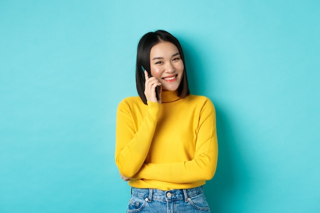 Giovane donna asiatica alla moda parlando al telefono, chiamando un amico e sorridente, in piedi con lo smartphone su sfondo blu.