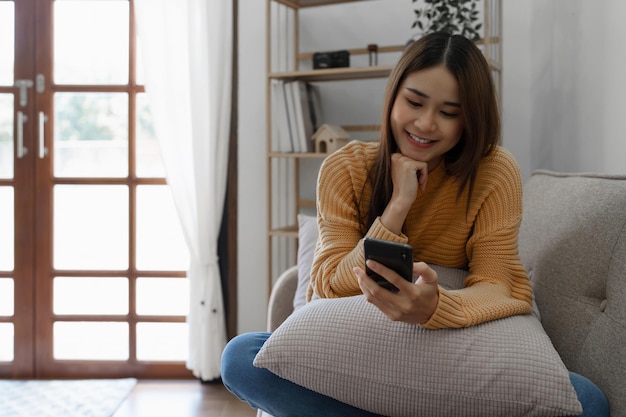 Giovane donna asiatica allegra che utilizza il telefono cellulare mentre era seduto su un divano a casa