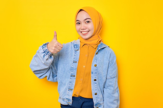 Giovane donna asiatica allegra in giacca di jeans che mostra i pollici in su gesto e guardando la fotocamera isolata su sfondo giallo