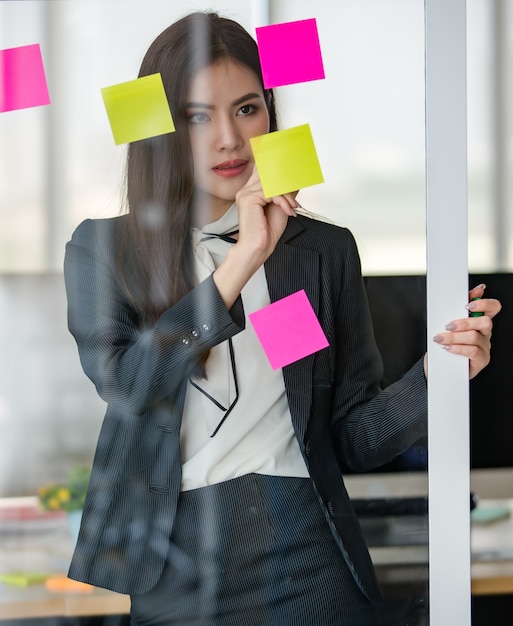 Giovane donna asiatica attraente in affari neri che legge e scrive su post-it su un pannello di vetro in un ufficio dall'aspetto moderno con sfondo sfocato delle finestre. Concetto per lo stile di vita moderno dell'ufficio.