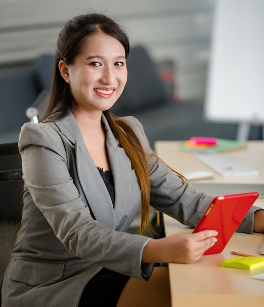 Giovane donna asiatica attraente in tailleur grigio che lavora su tablet rosso in un ufficio dall'aspetto moderno con sfondo sfocato delle finestre. Concetto per lo stile di vita moderno dell'ufficio.