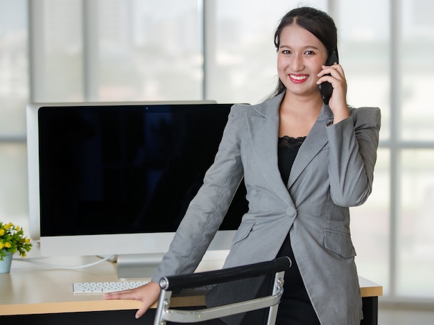 Giovane donna asiatica attraente in tailleur grigio che parla al telefono cellulare in un ufficio dall'aspetto moderno con sfondo sfocato delle finestre. Concetto per lo stile di vita moderno dell'ufficio.