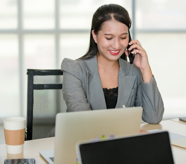 Giovane donna asiatica attraente in tailleur grigio seduta a parlare al telefono cellulare in un ufficio dall'aspetto moderno con sfondo sfocato delle finestre. Concetto per lo stile di vita moderno dell'ufficio.