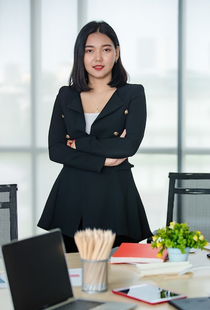 Giovane donna asiatica attraente in tailleur nero in piedi braccio incrociato con fiducia in un ufficio dall'aspetto moderno. Concetto per lo stile di vita moderno dell'ufficio.
