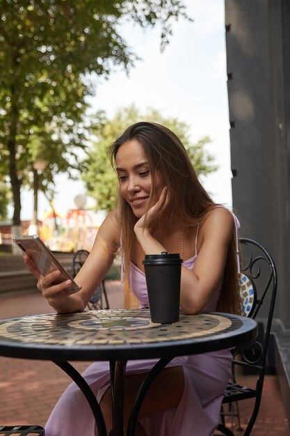 Giovane donna asiatica ben vestita seduta al tavolo con il caffè, utilizzando lo smartphone