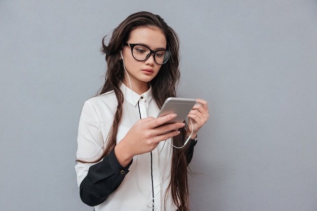 Giovane donna asiatica carina con gli occhiali che ascolta musica sul telefono isolato su grigio