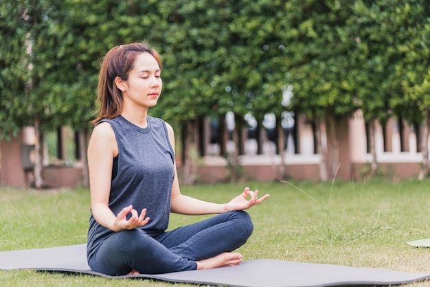 Giovane donna asiatica che fa yoga all'aperto nella posa meditata del loto seduta sull'erba verde con gli occhi chiusi al parco giardino, concetto di assistenza sanitaria