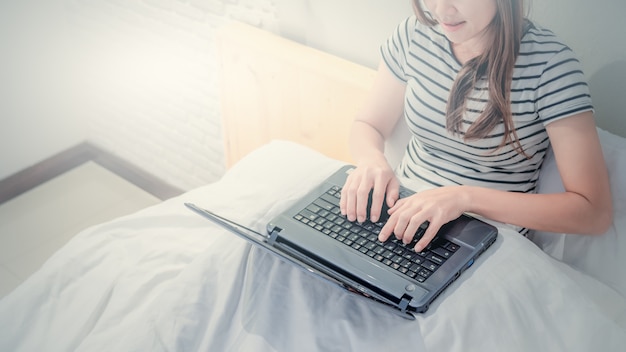 Giovane donna asiatica che gioca con il computer portatile sul suo letto in camera da letto bianca.