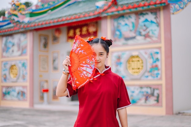 Giovane donna asiatica che indossa un abito cheongsam qipao con ventaglio pieghevole cinese su sfondo rosso per Chine