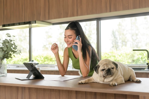 Giovane donna asiatica che lavora con il tablet e si diverte con il suo cane in cucina a casa
