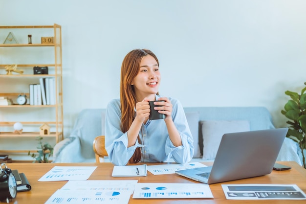 Giovane donna asiatica che lavora da casa usando il computer e bevendo caffè nella sua stanza documenta la finanza e la conferenza online per nuovi progetti