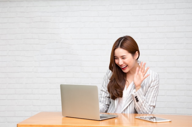 Giovane donna asiatica che parla esaminando computer portatile e che sorride mentre lavorando a casa, videoconferenza o concetto di addestramento online.