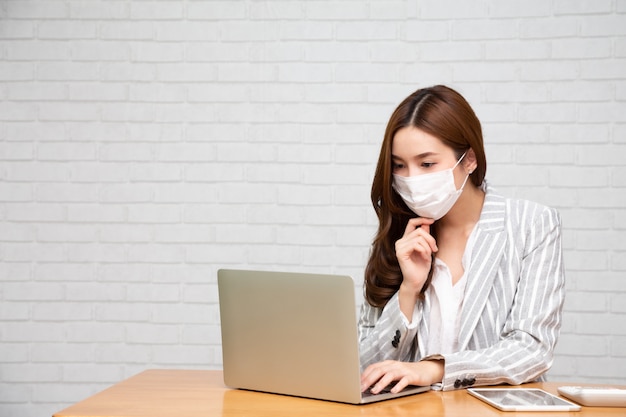 Giovane donna asiatica che per mezzo del computer portatile e indossando la maschera di protezione.