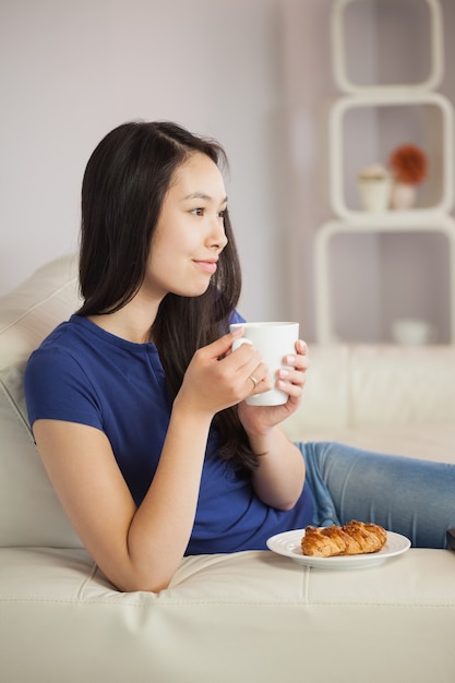 Giovane donna asiatica che si siede sul sofà che mangia caffè withpastry