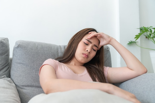 Giovane donna asiatica che si tocca la testa sentendo mal di testa mentre è seduta sul divano in soggiorno.