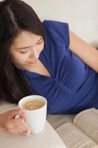 Giovane donna asiatica che si trova sul sofà che legge un libro che tiene il suo caffè