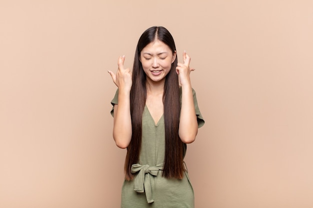 Giovane donna asiatica che sorride e incrociando ansiosamente entrambe le dita, sentendosi preoccupata e desiderando o sperando in buona fortuna