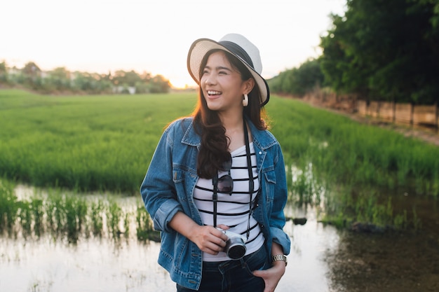 Giovane donna asiatica che sorride in cappello con la macchina fotografica. Ragazza che gode della bellissima natura con il tramonto.