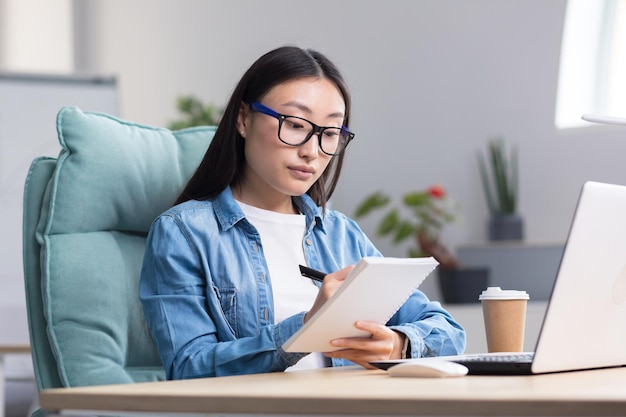 Giovane donna asiatica che studia online mentre è seduto in ufficio a scrivere appunti su un notebook utilizzando il laptop per studiare