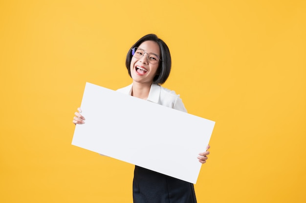 Giovane donna asiatica che tiene carta in bianco con il fronte sorridente e che guarda sulla parete arancio per i cartelli pubblicitari.