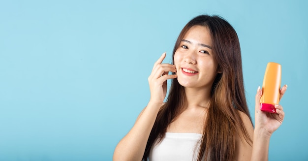 Giovane donna asiatica che tiene in mano la lozione solare con la mano che tocca il viso scatto in studio isolato su sfondo blu felice sorridente adolescente donna presente protezione solare cura della pelle