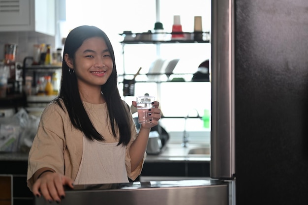 Giovane donna asiatica che tiene in mano un bicchiere d'acqua e sorride alla telecamera mentre si trova in cucina.