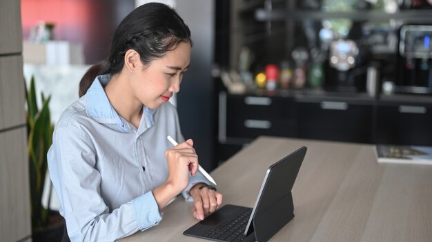 Giovane donna asiatica che tiene la penna stilo e lavora online con il tablet del computer al bancone della cucina di casa.