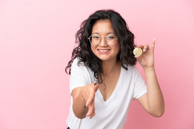 Giovane donna asiatica che tiene un Bitcoin isolato su sfondo rosa che stringe la mano per aver chiuso un buon affare