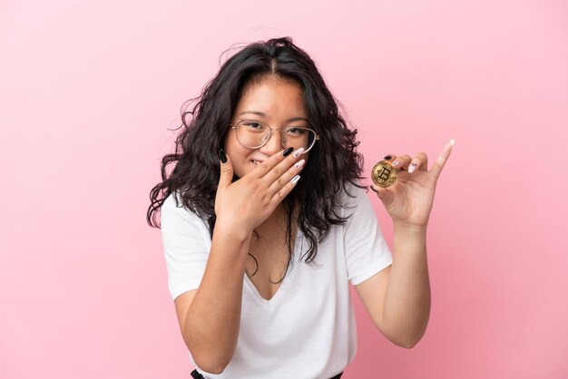 Giovane donna asiatica che tiene un Bitcoin isolato su sfondo rosa felice e sorridente che copre la bocca con la mano