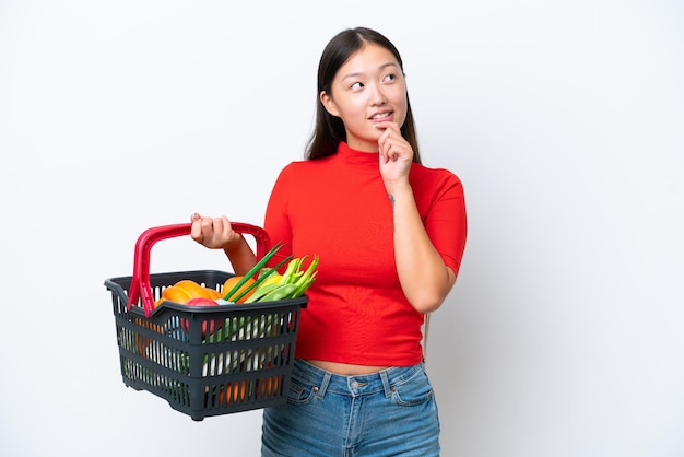 Giovane donna asiatica che tiene un carrello pieno di cibo isolato su sfondo bianco e guardando in alto