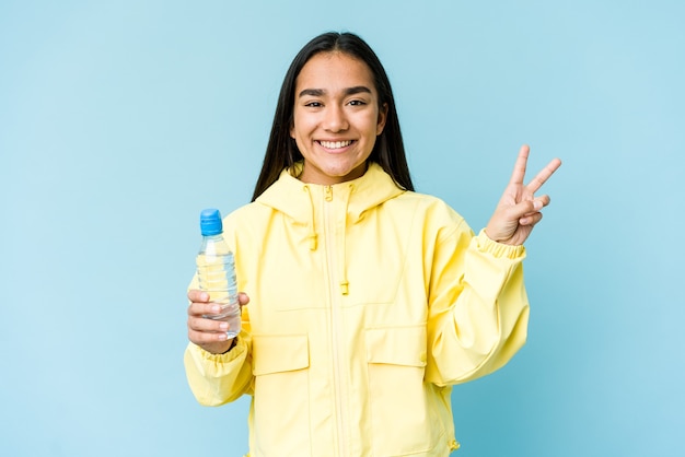 Giovane donna asiatica che tiene una bottiglia di acqua isolata sull'azzurro gioioso e spensierato che mostra un simbolo di pace con le dita.