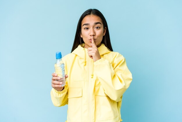 Giovane donna asiatica che tiene una bottiglia di acqua isolata sulla parete blu che mantiene un segreto o che chiede il silenzio.