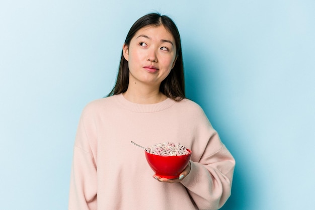 Giovane donna asiatica che tiene una ciotola di cereali isolata su sfondo blu che sogna di raggiungere obiettivi e scopi