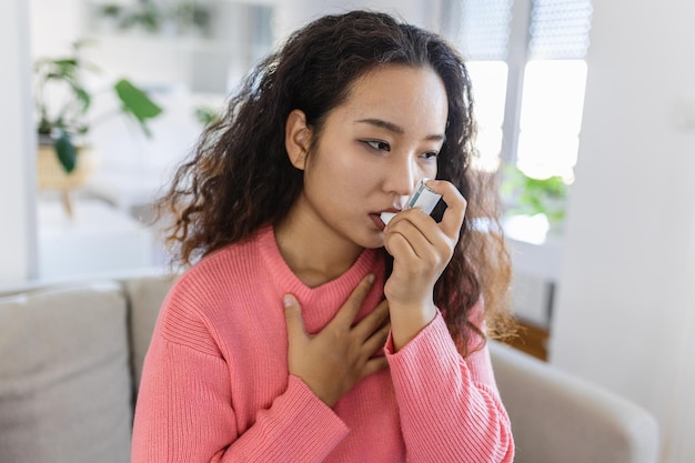 Giovane donna asiatica che usa l'inalatore per l'asma sul divano a casa nel soggiorno