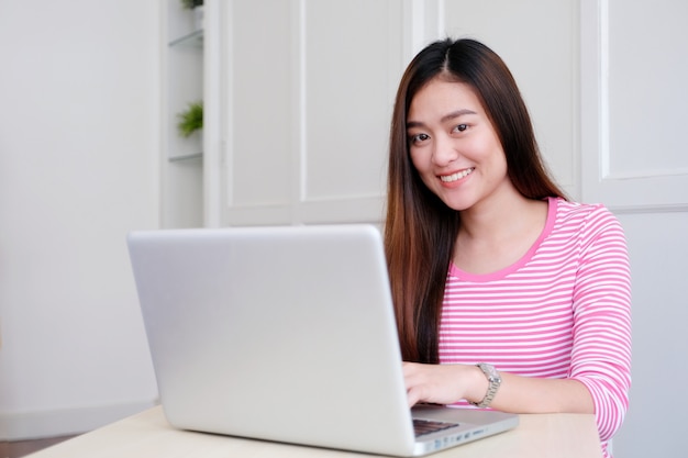 Giovane donna asiatica che utilizza computer portatile che si siede nel fondo della stanza bianca