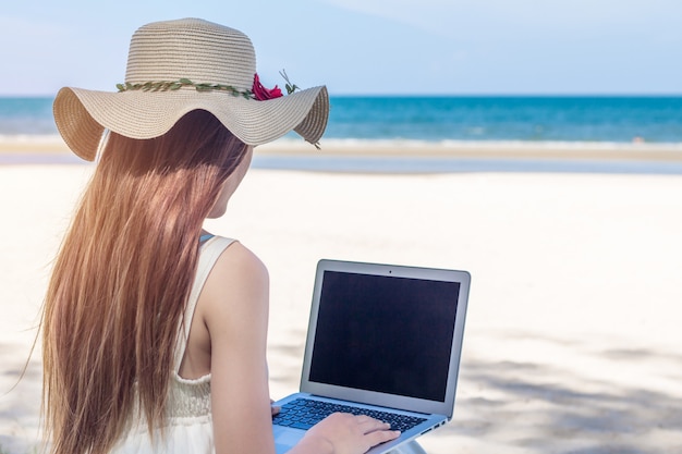 Giovane donna asiatica che utilizza computer portatile in vestito che si siede sulla spiaggia, Freelancer della ragazza che lavorano