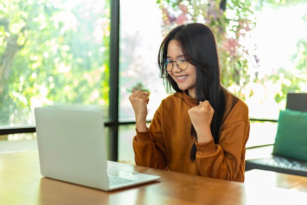 Giovane donna asiatica che utilizza il computer portatile e alza le braccia di gioia con estrema gioia vincitore del premio speciale